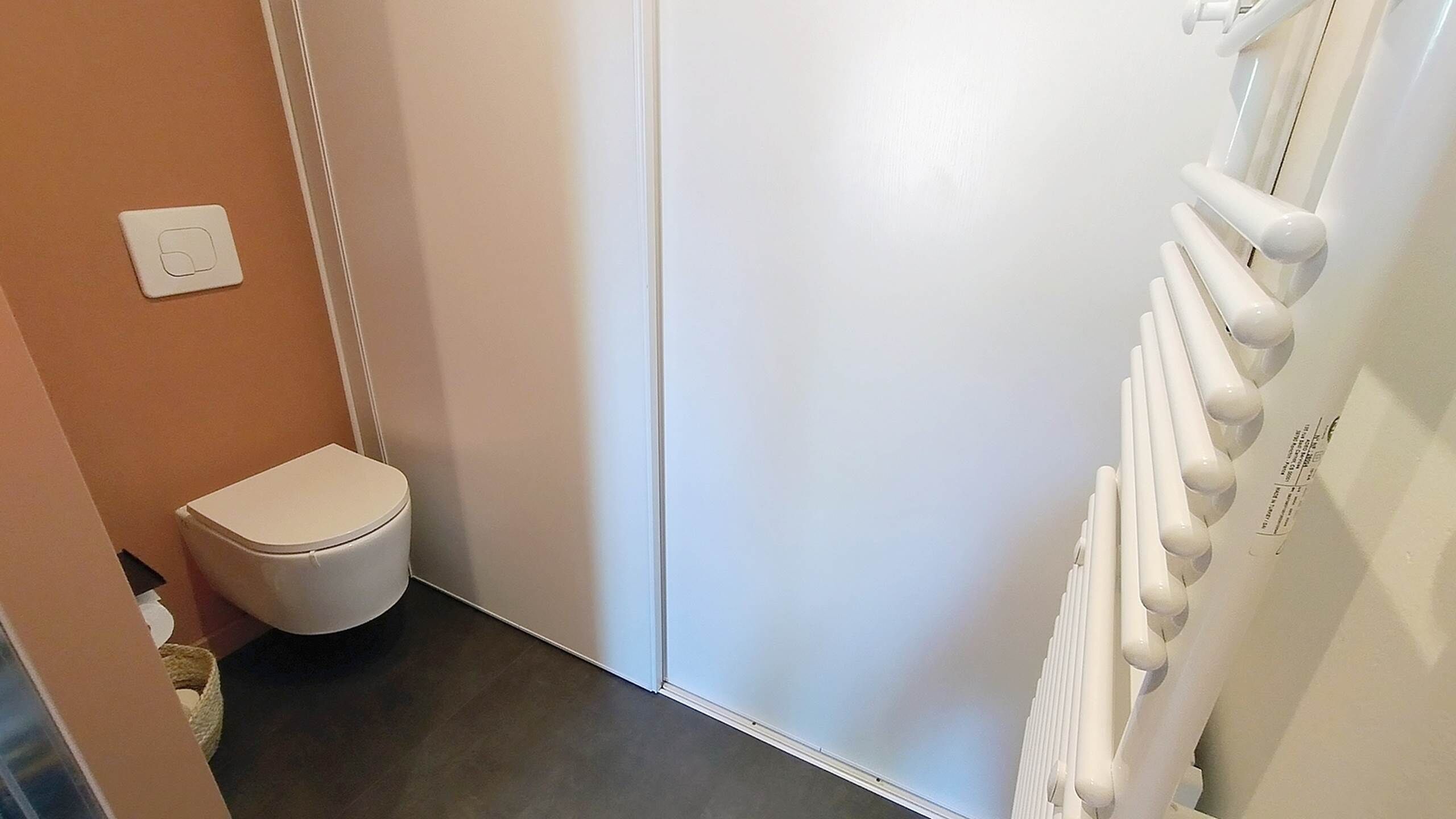 Une petite salle de bain étroite, avec des toilettes blanches, un radiateur blanc et un interrupteur sur le mur.
