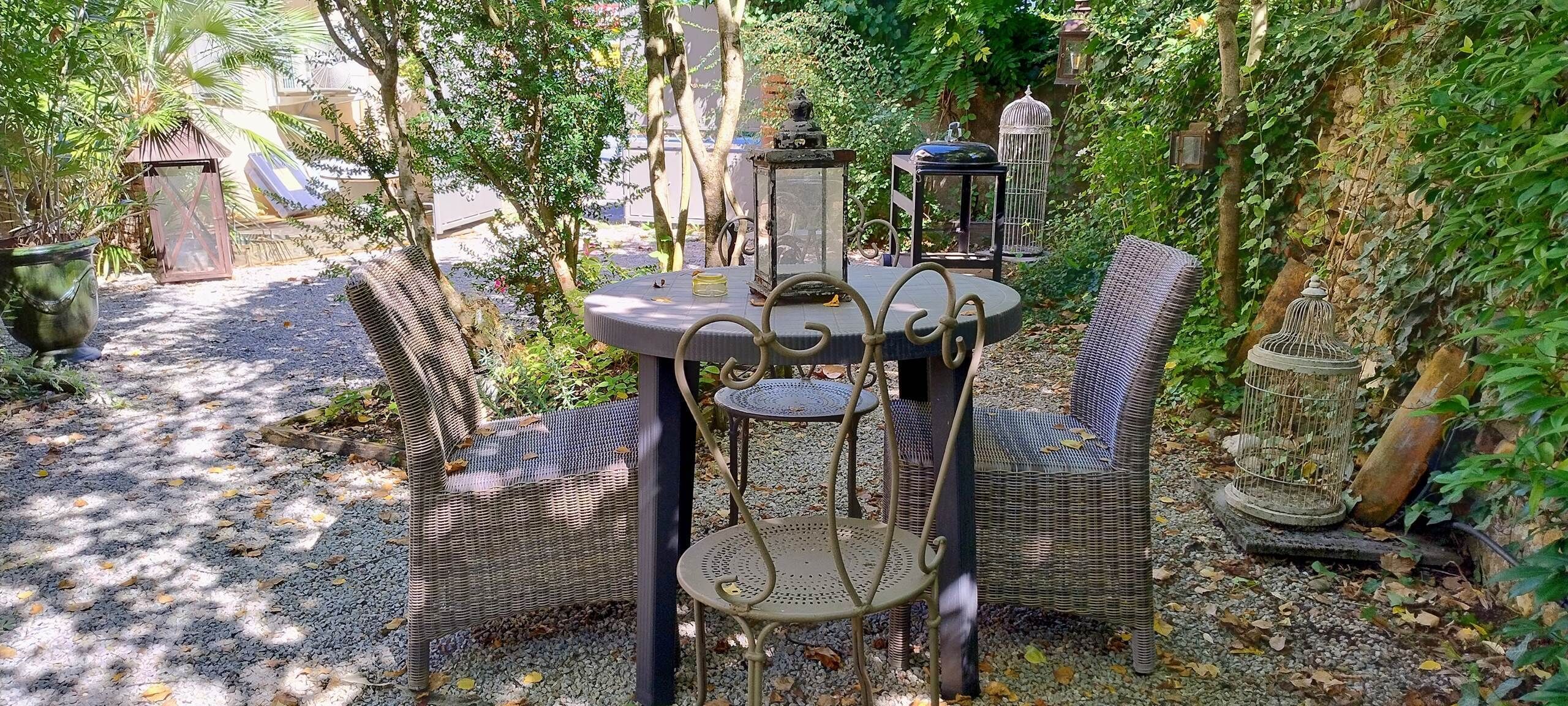 An outdoor seating area with wicker furniture and a round table, surrounded by trees and plants.