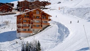 Een meerlaags houten gebouw met balkons, omringd door besneeuwde hellingen en skipistes in de verte.