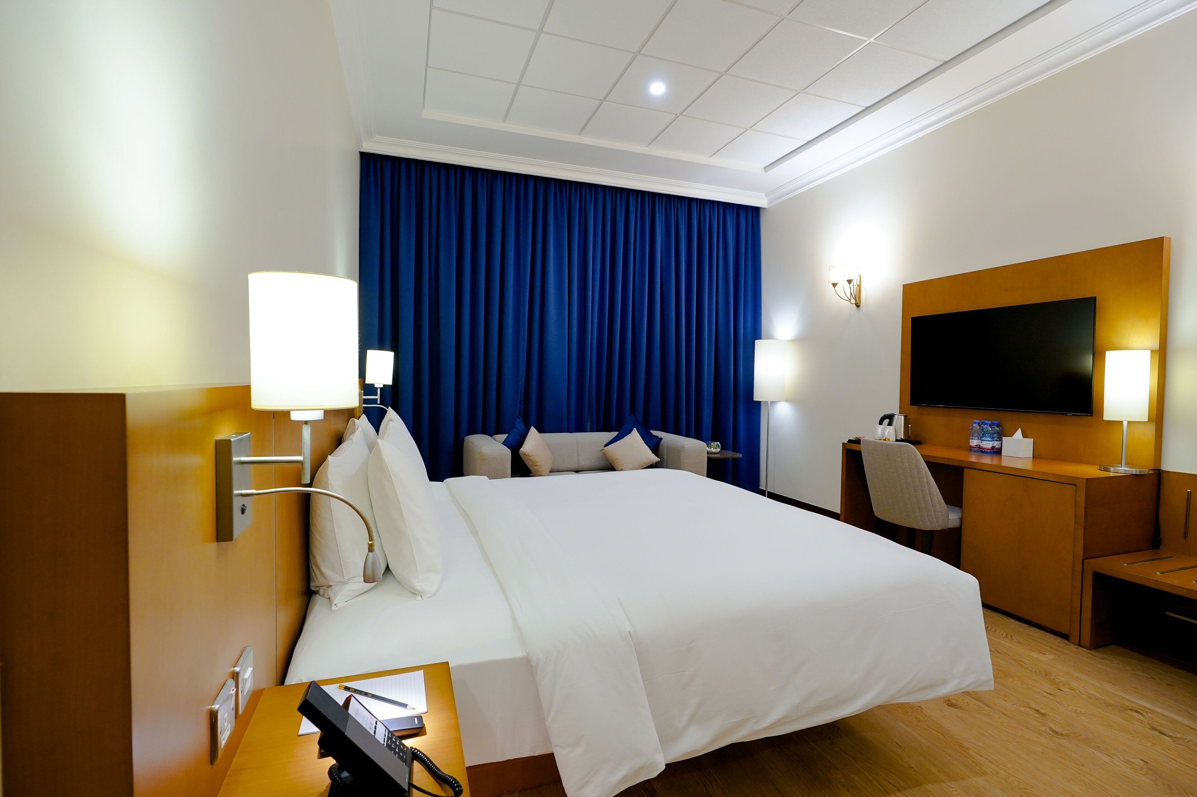 A hotel room with a large bed, a desk with a telephone, a flat-screen TV, and a desk lamp.