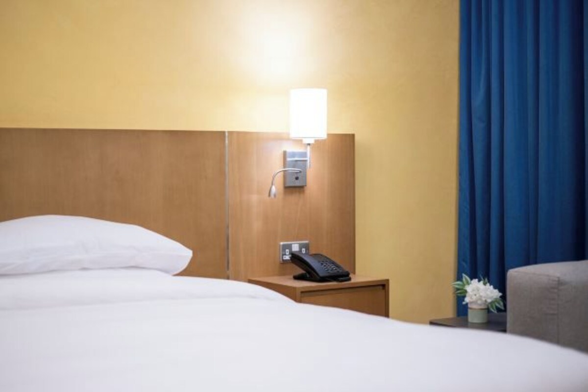 Una habitación de hotel con cama, teléfono en la mesita de noche y lámpara fijada a la pared.