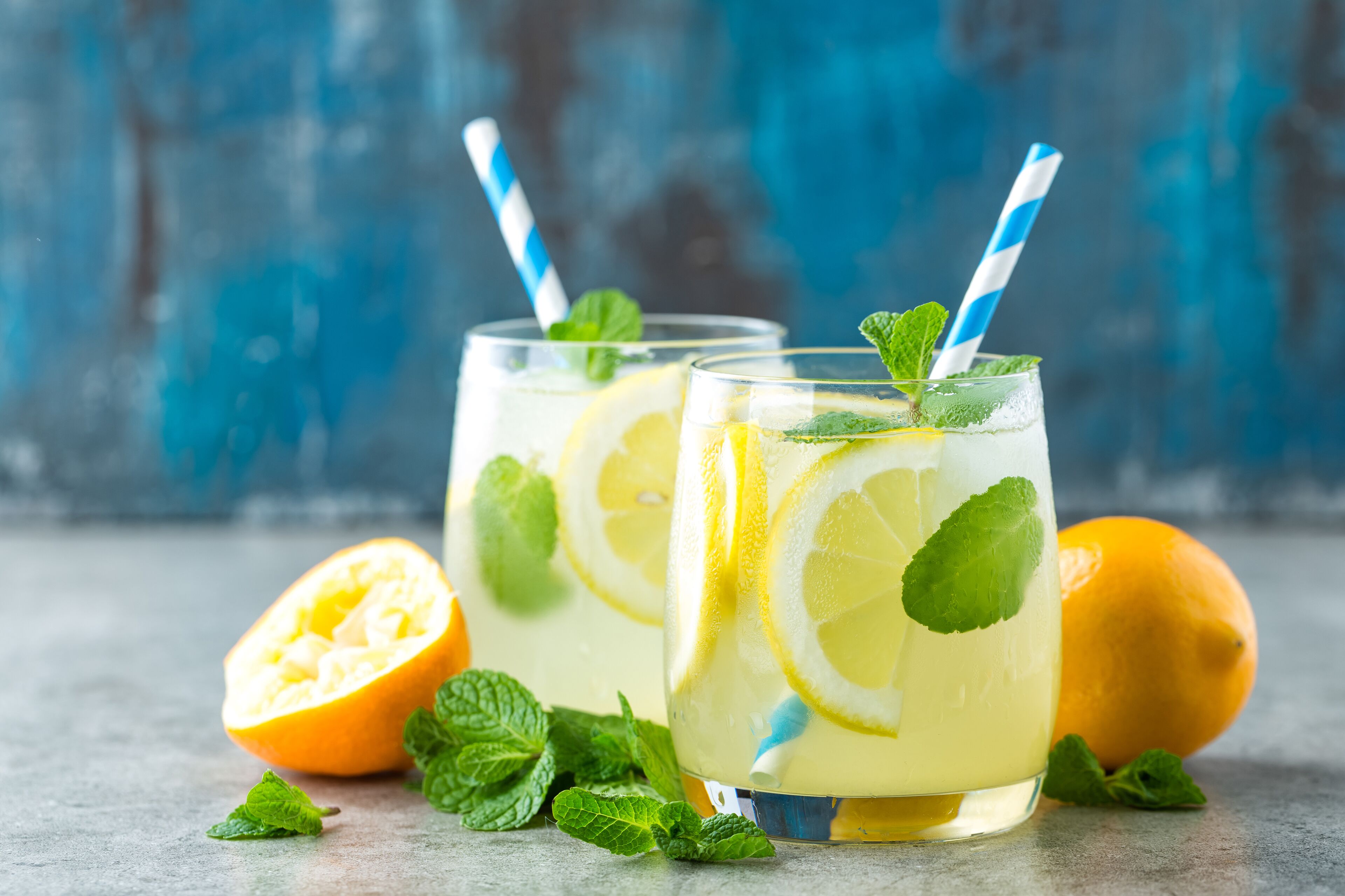 Two glasses of lemonade with lemon slices and mint leaves, a halved lemon, and mint leaves on a textured surface.