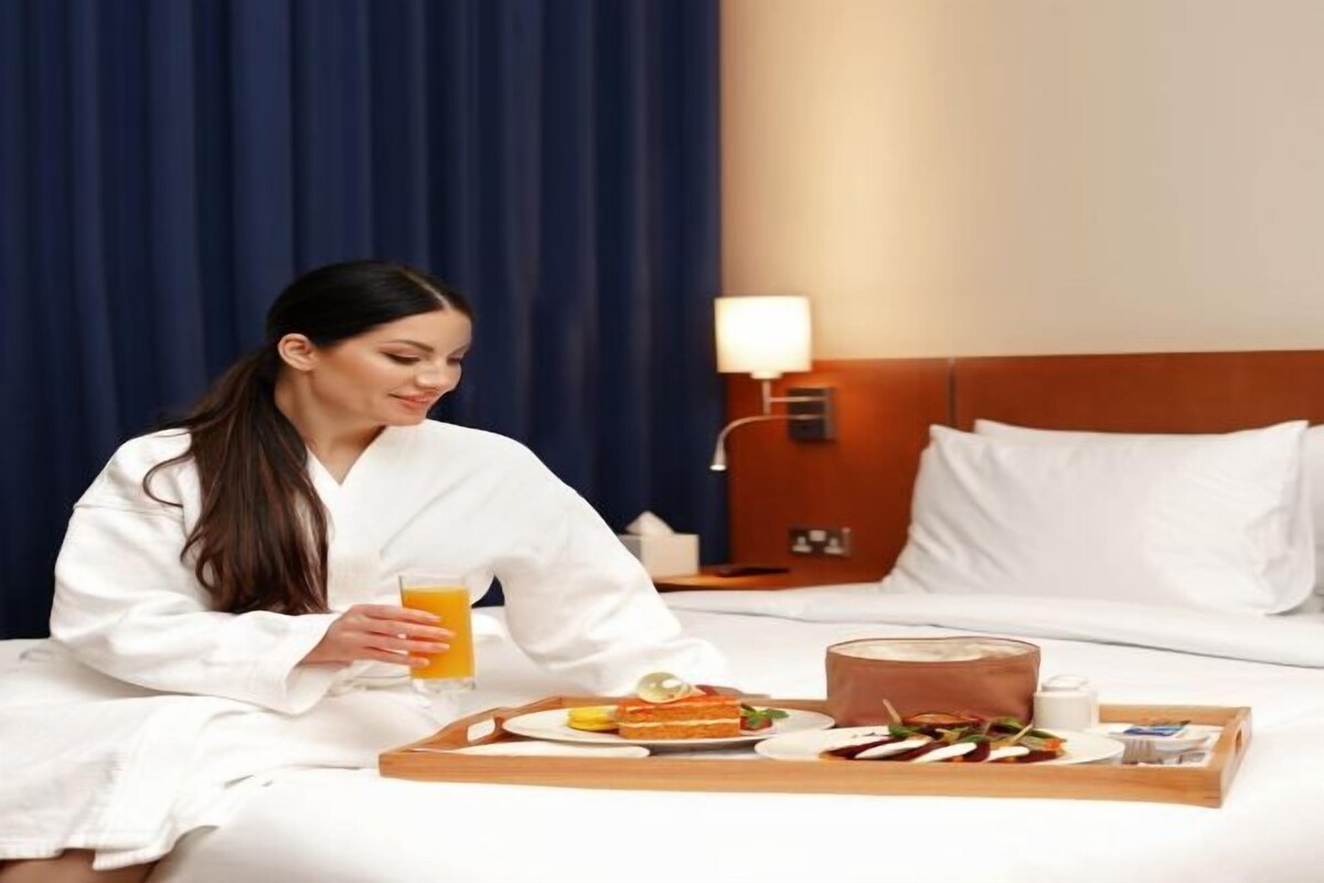 Una habitación de hotel con una cama, una mesita de noche con lámpara y una bandeja de desayuno con comida y un vaso de jugo de naranja.
