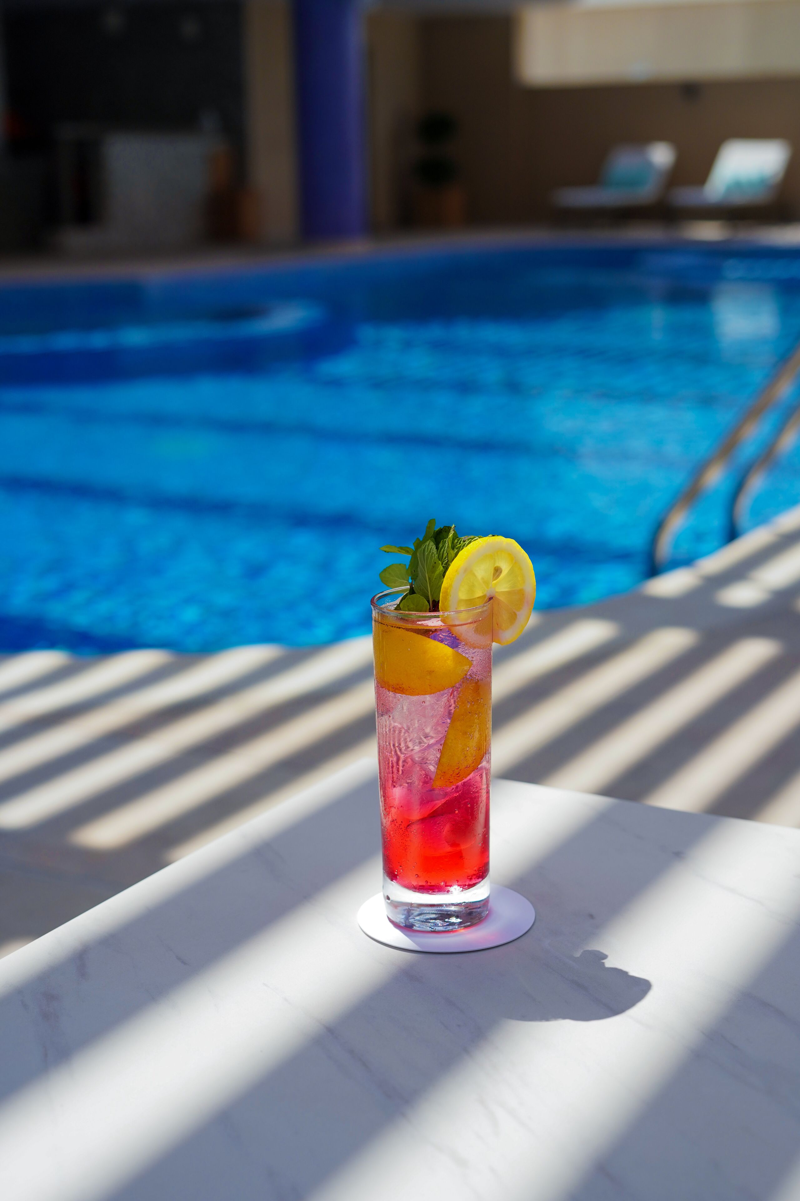 A glass of red drink with lemon and mint on a white table by a pool.
