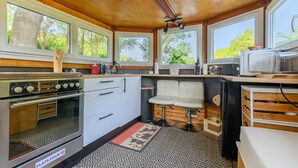 A compact kitchen with a stove, refrigerator, and microwave. There are large windows providing natural light and views of greenery outside.