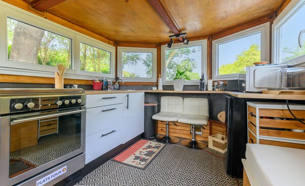 A compact kitchen with a stove, refrigerator, and microwave. There are large windows providing natural light and views of greenery outside.