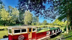 A narrowboat docked at a riverside location with a grassy bank and trees in the background.