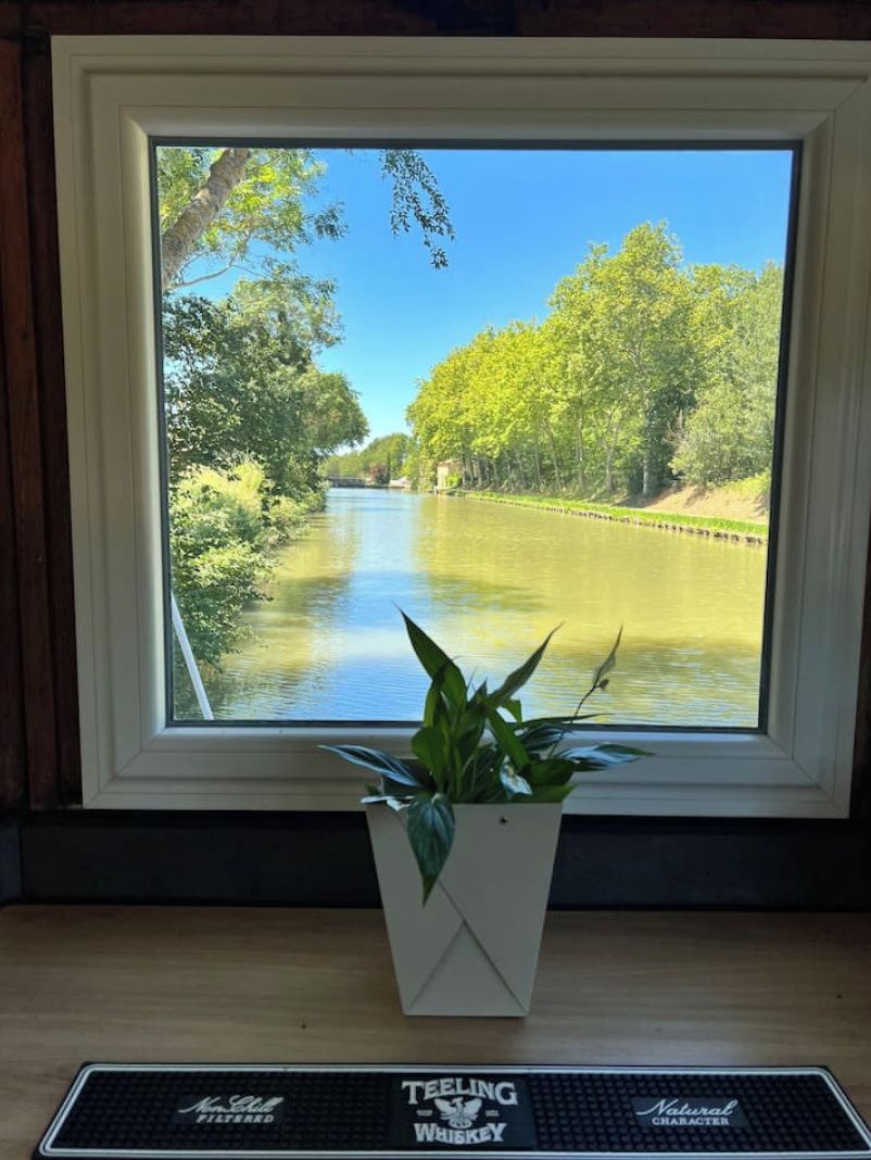 Een raam met uitzicht op een rivier, een bloempot en een toetsenbord op een bureau.