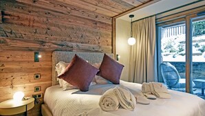 Een slaapkamer met een houten plafond, een bed met witte beddengoed en twee kussens, een nachtkastje met een lamp, en een besneeuwde tuin zichtbaar door een groot raam.