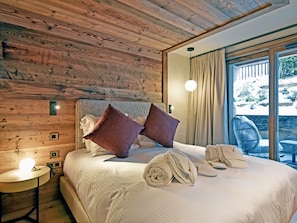 A bedroom with a wooden ceiling, a bed with white bedding and two pillows, a bedside table with a lamp, and a view of a snowy garden through a large window.