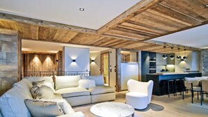 En moderne stue med sofa, lænestole og sofabord. Der er et køkkenområde med en bar og barstole. Rummet har synlige træbjælker og stenvægge.