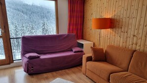 Una sala de estar con un sofá morado, un sofá marrón, un pequeño banquito y una pared de madera con una ventana que da a un bosque nevado.