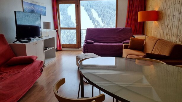 Una sala de estar moderna con una mesa de comedor de vidrio, sofás rojos y marrones, un televisor sobre un mueble blanco y vistas a montañas nevadas a través de amplios ventanales.