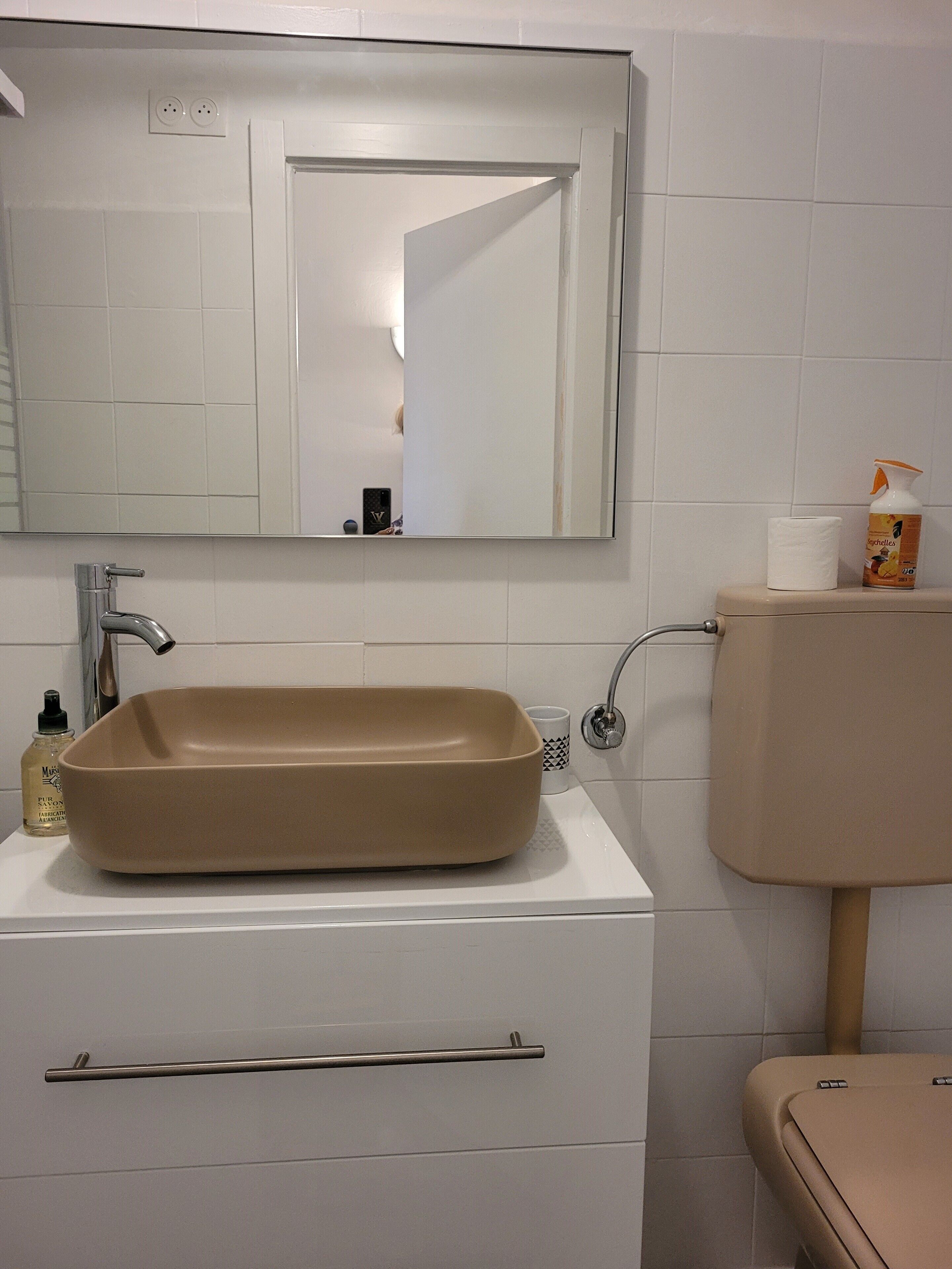 A bathroom with a beige sink, a mirror, a toilet, and a bottle of hand soap.