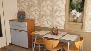 A dining area with a glass table, four yellow chairs, a small cabinet, and a wall with a palm leaf pattern.
