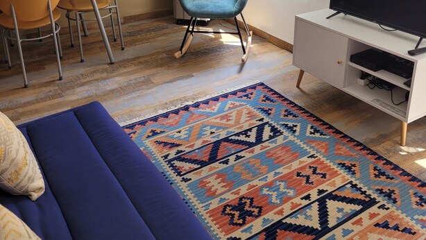 A living room with a sofa, a TV stand, a patterned rug, a dining area with chairs, a blue armchair, and a wall with a leaf pattern.