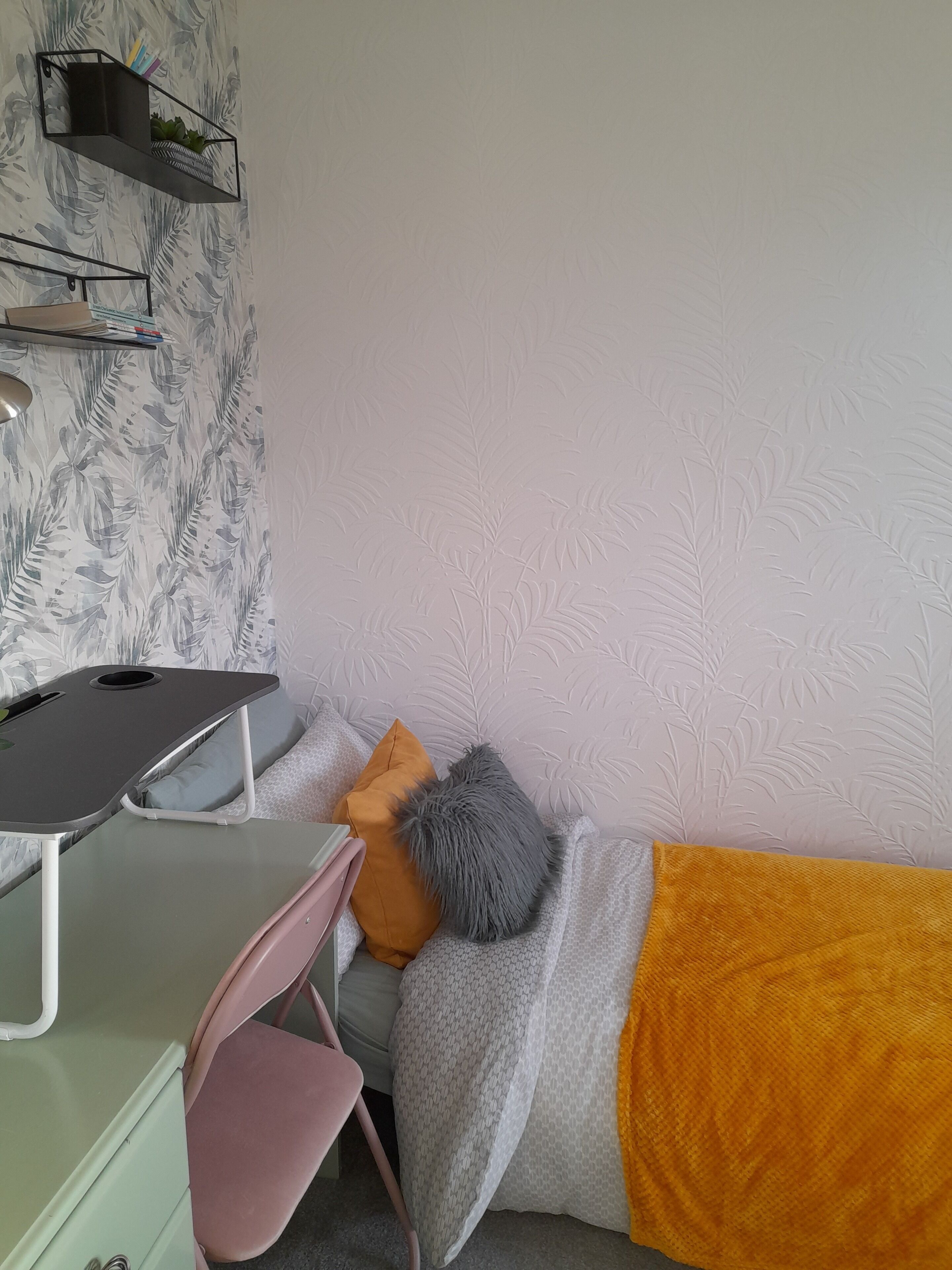 A bedroom with a bed, a desk, and a chair. There is a shelf with books and a plant.