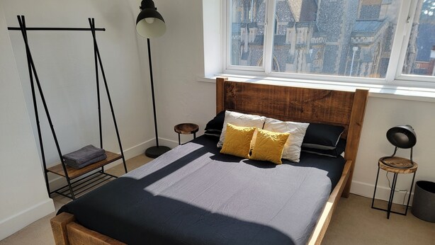 A bedroom with a wooden bed frame, a black and white bedding set, a standing floor lamp, and a view of a building through a window. - NEW! Rustic Loft Escape Bournemouth (5mins to Beach, Uni, Bars, Shops) Park FREE (Bournemouth)