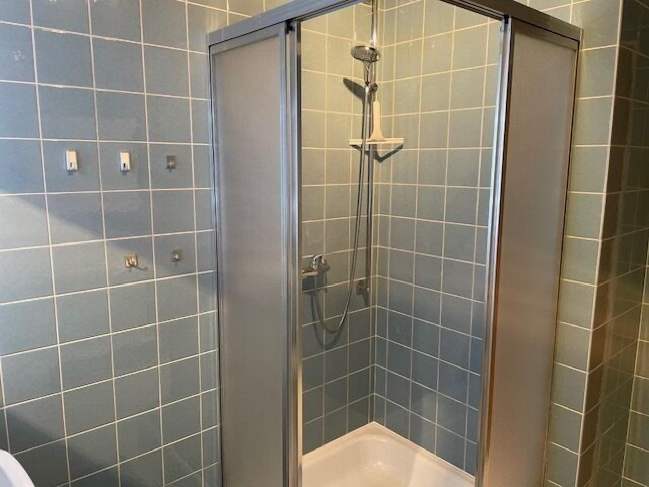 Eine Dusche mit Glastür, Duschbrausehandstück und am Wand montiertem Wasserhahn.