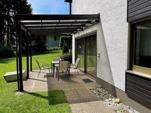 Une terrasse couverte avec une table et des chaises, une pelouse et un bâtiment à l’extérieur blanc et doté de grandes fenêtres.