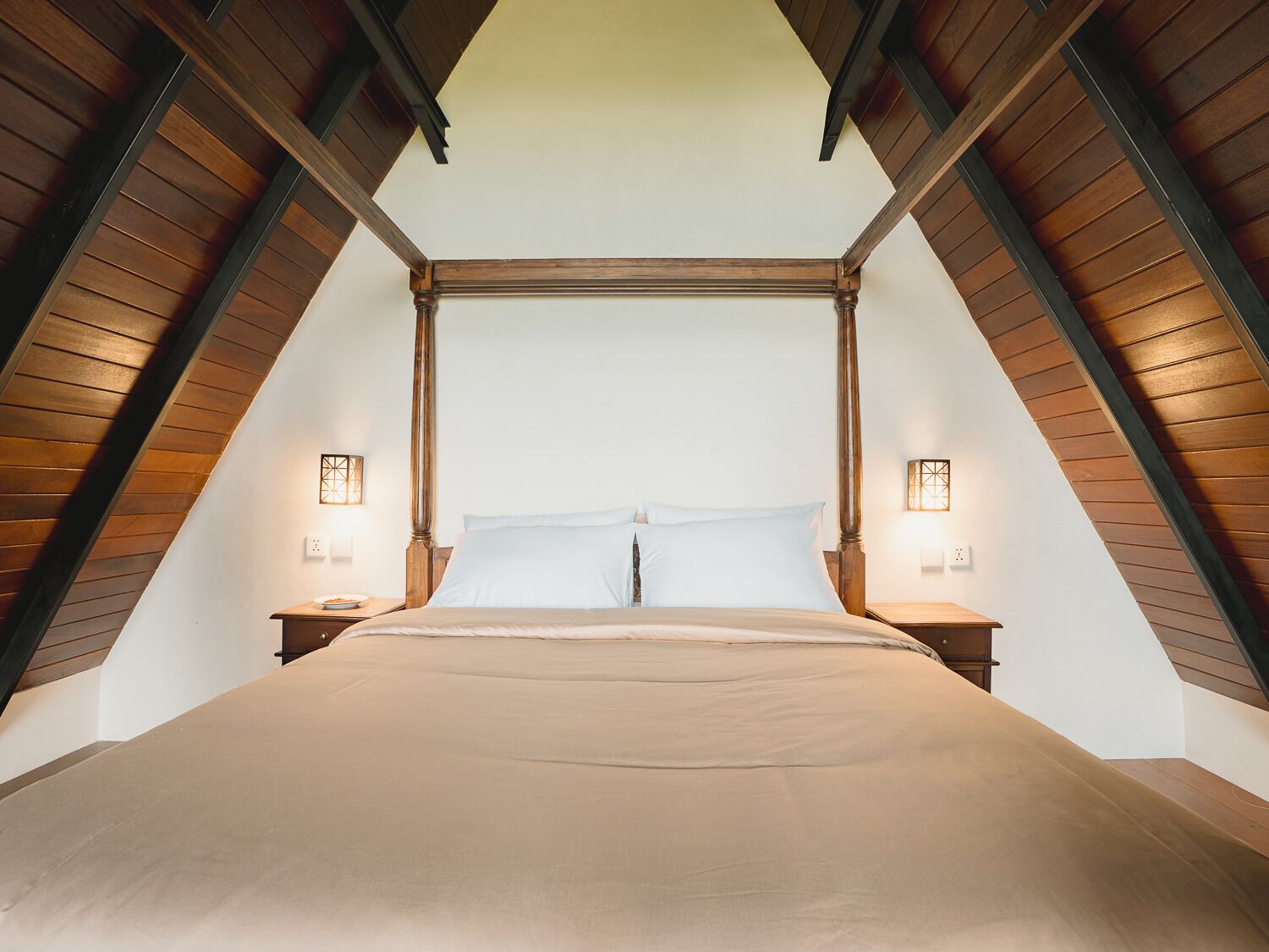 A high-ceilinged room with a large bed centered under a wooden beam structure, flanked by two bedside tables with lamps.