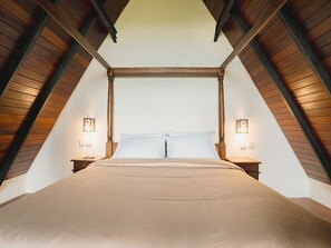 Ein Zimmer mit hohen Decken, ein großes Bett unter einer Holzbalkenkonstruktion, flankiert von zwei Nachttischen mit Lampen.
