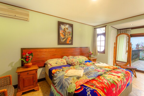 A bedroom with a wooden headboard, a bed with a colorful floral bedspread, a nightstand with a vase of flowers, and a large mirror. - Pacung Indah Hotel & Restaurant (Baturiti)