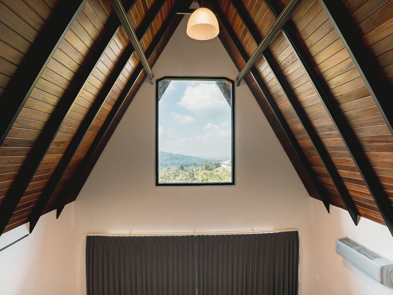 Una habitación con techo inclinado, una ventana que ofrece vistas al exterior, una lámpara en el techo y un equipo de aire acondicionado.