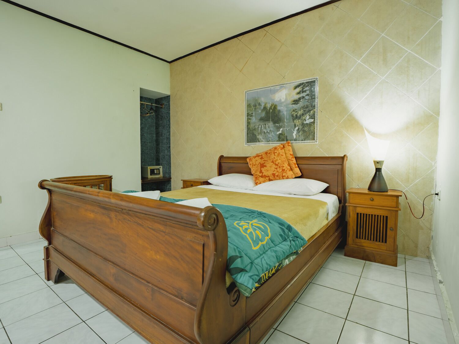Un dormitorio con una cama de madera, una mesita de noche con lámpara, un cuadro en la pared y suelo de baldosas.