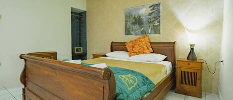 A bedroom with a wooden bed, a nightstand with a lamp, a wall-mounted painting, and a tiled floor.
