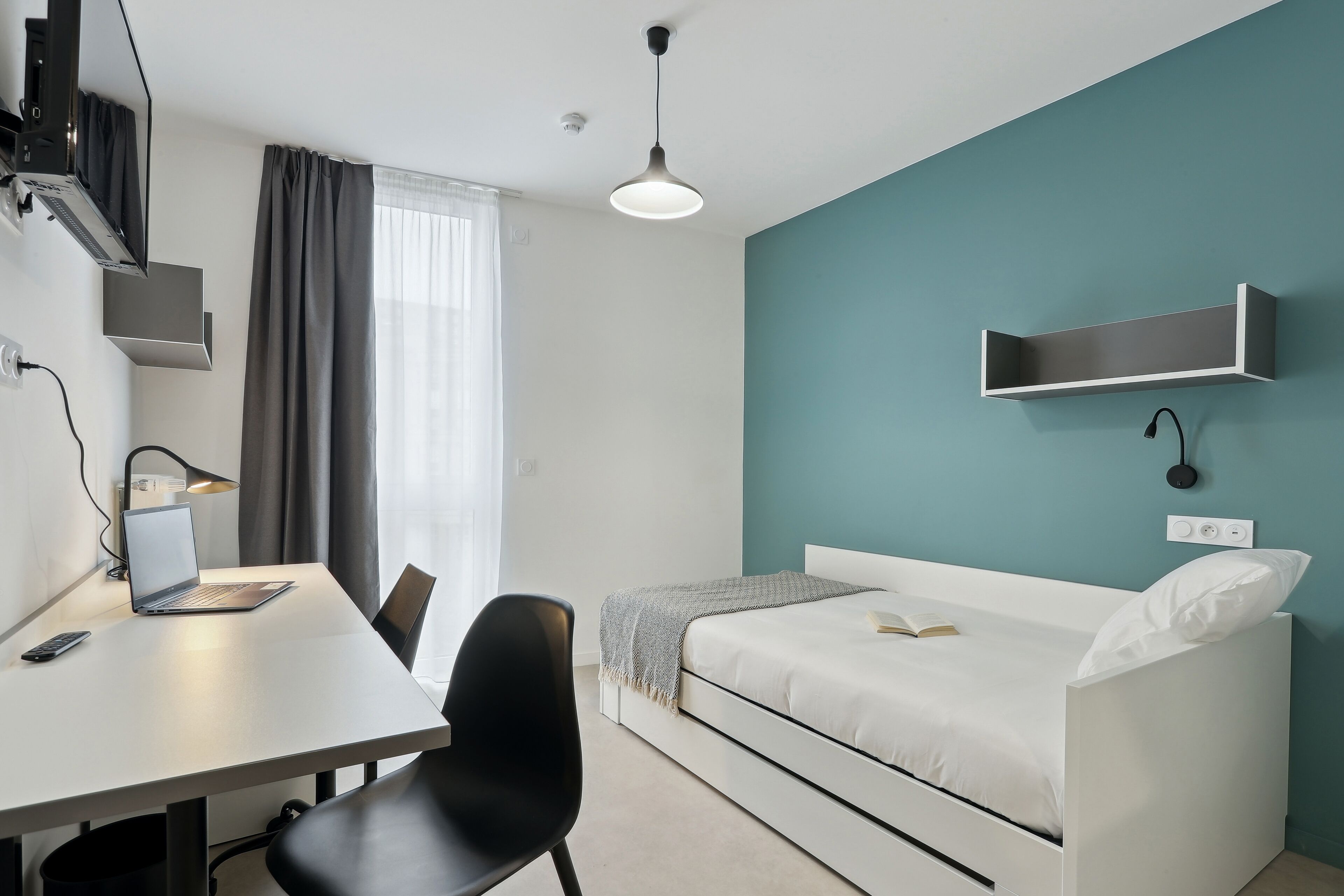 A modern hotel room with a white desk, laptop, and chair, a white bed with a teal accent wall, and a pendant light.