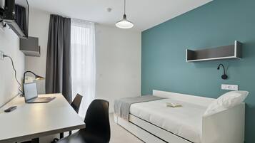 A modern hotel room with a white desk, laptop, and chair, a white bed with a teal accent wall, and a pendant light.