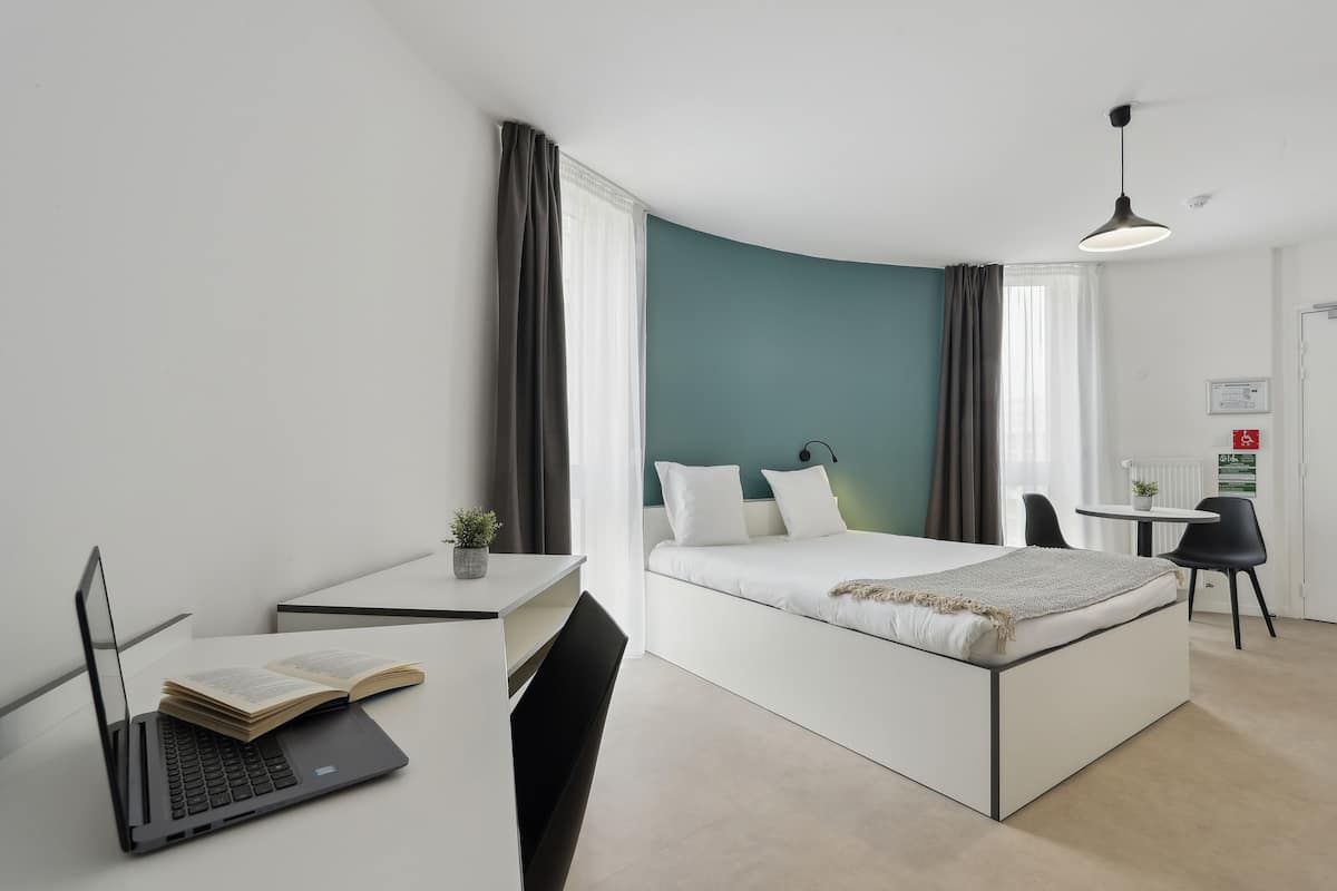 Een moderne slaapkamer met een bed, een bureau met een laptop en een open boek, een kleine tafel met een plant en een stoel.