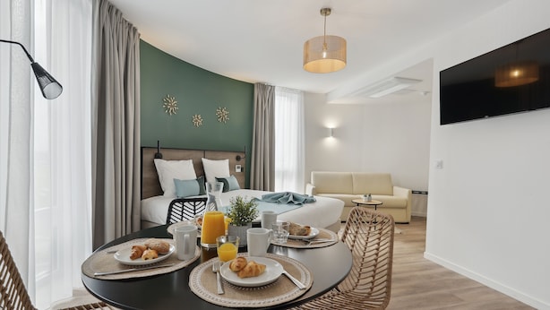 A modern hotel room with a dining area, breakfast setup, a bed with a view, and a TV mounted on the wall. - All Suites Appart Hôtel | Noisy-le-Grand (Noisy-le-Grand)