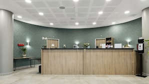 A reception area with a curved wooden counter, a potted plant, a desk with a computer, and a coffee machine. - All Suites Appart Hôtel | Noisy-le-Grand (Noisy-le-Grand)