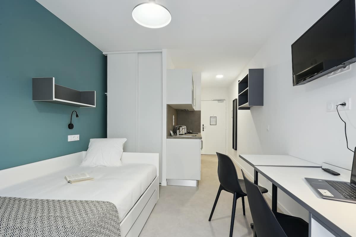 Een moderne hotelkamer met een bed, een bureau met een laptop, een televisie en een kitchenette op de achtergrond.