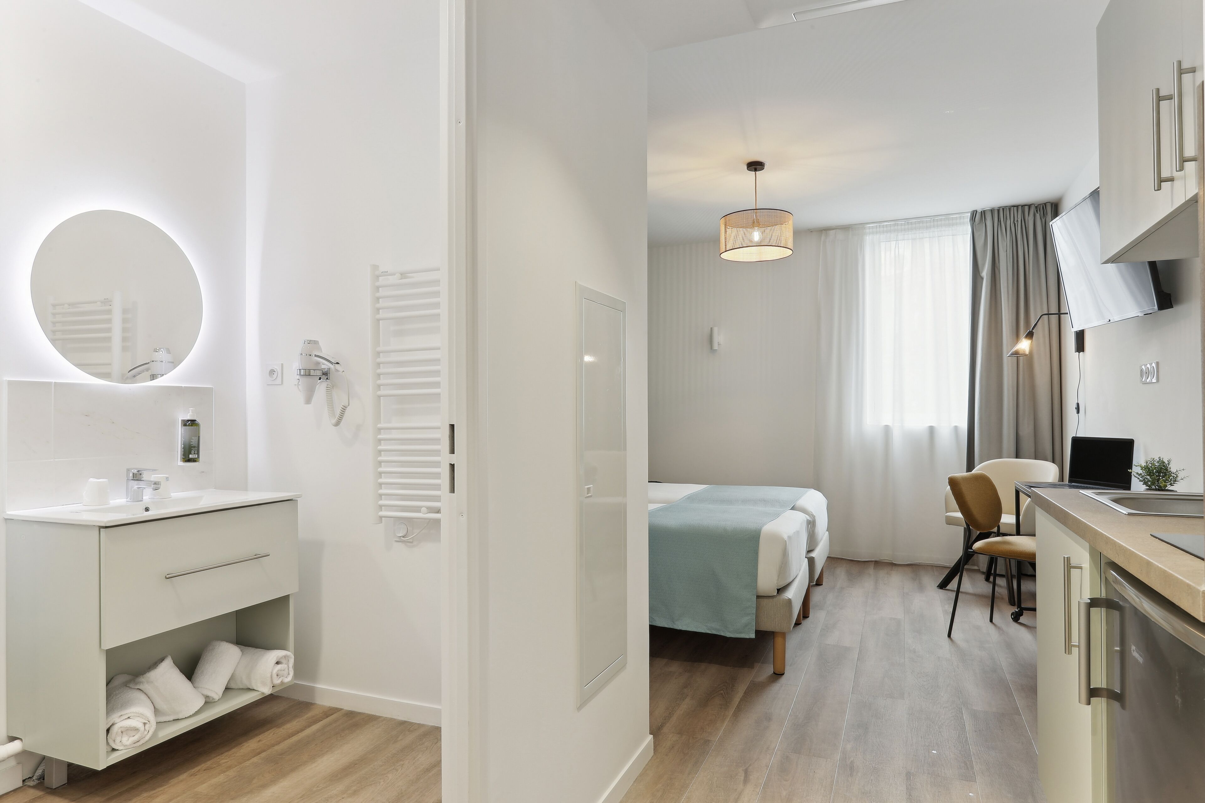 Une chambre d’hôtel moderne avec une coiffeuse blanche, un miroir rond, un lit recouvert d’une courtepointe bleue, un bureau avec un ordinateur et une chaise.