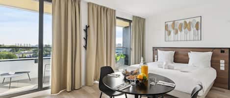 A modern hotel room with a large bed, a dining table with breakfast items, and a balcony with a view of the city.