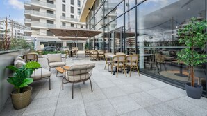 An outdoor seating area with wicker furniture and potted plants, overlooking a modern building with large windows. - All Suites Appart Hôtel | Paris Ouest - Colombes (Colombes)