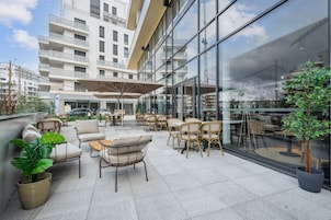 Un espacio al aire libre con mobiliario de mimbre y plantas en macetas, con vistas a un edificio moderno con grandes ventanales.
