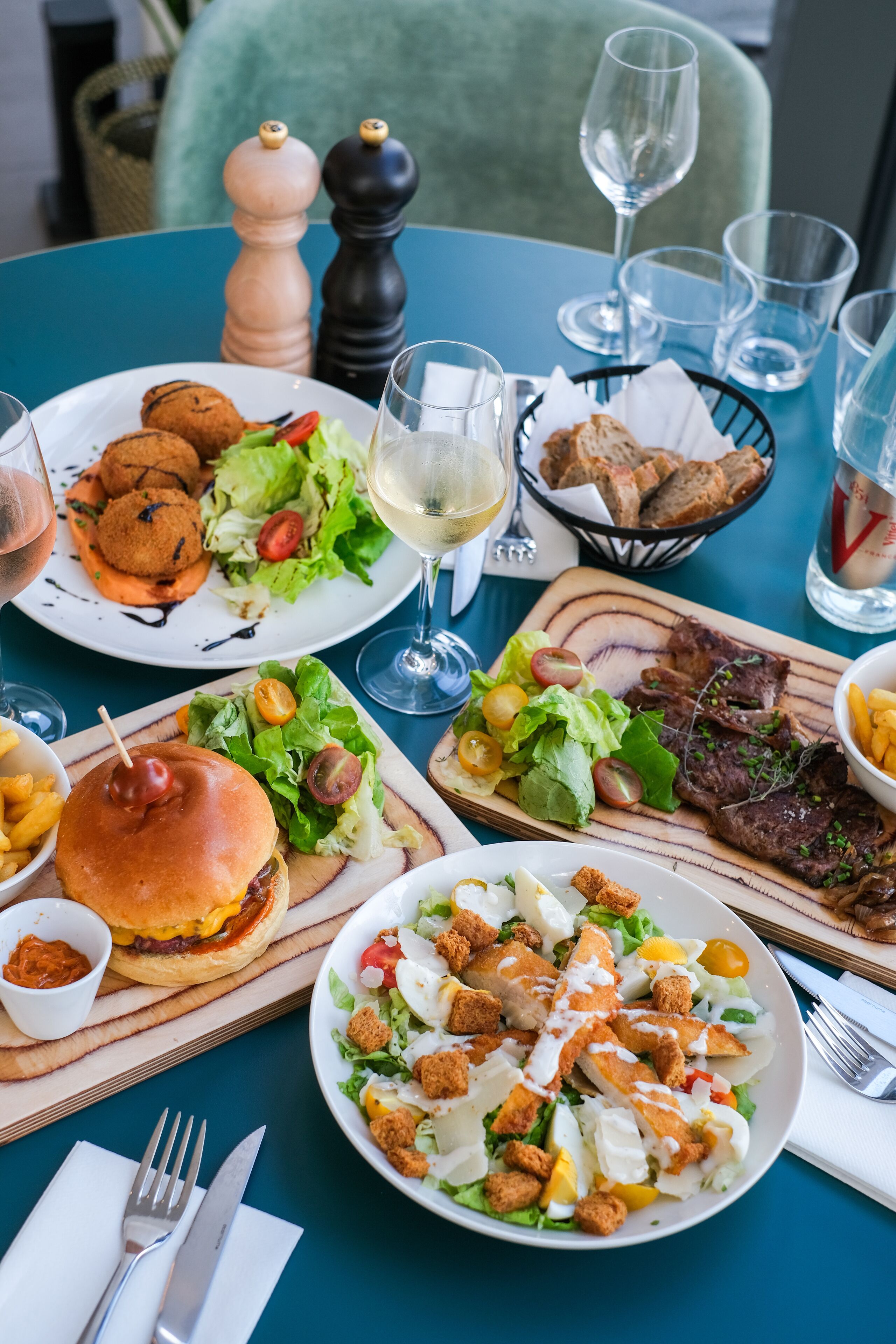 Mesa posta com diversos pratos, incluindo hambúrgueres, saladas e bife, acompanhada de copos de vinho e talheres.