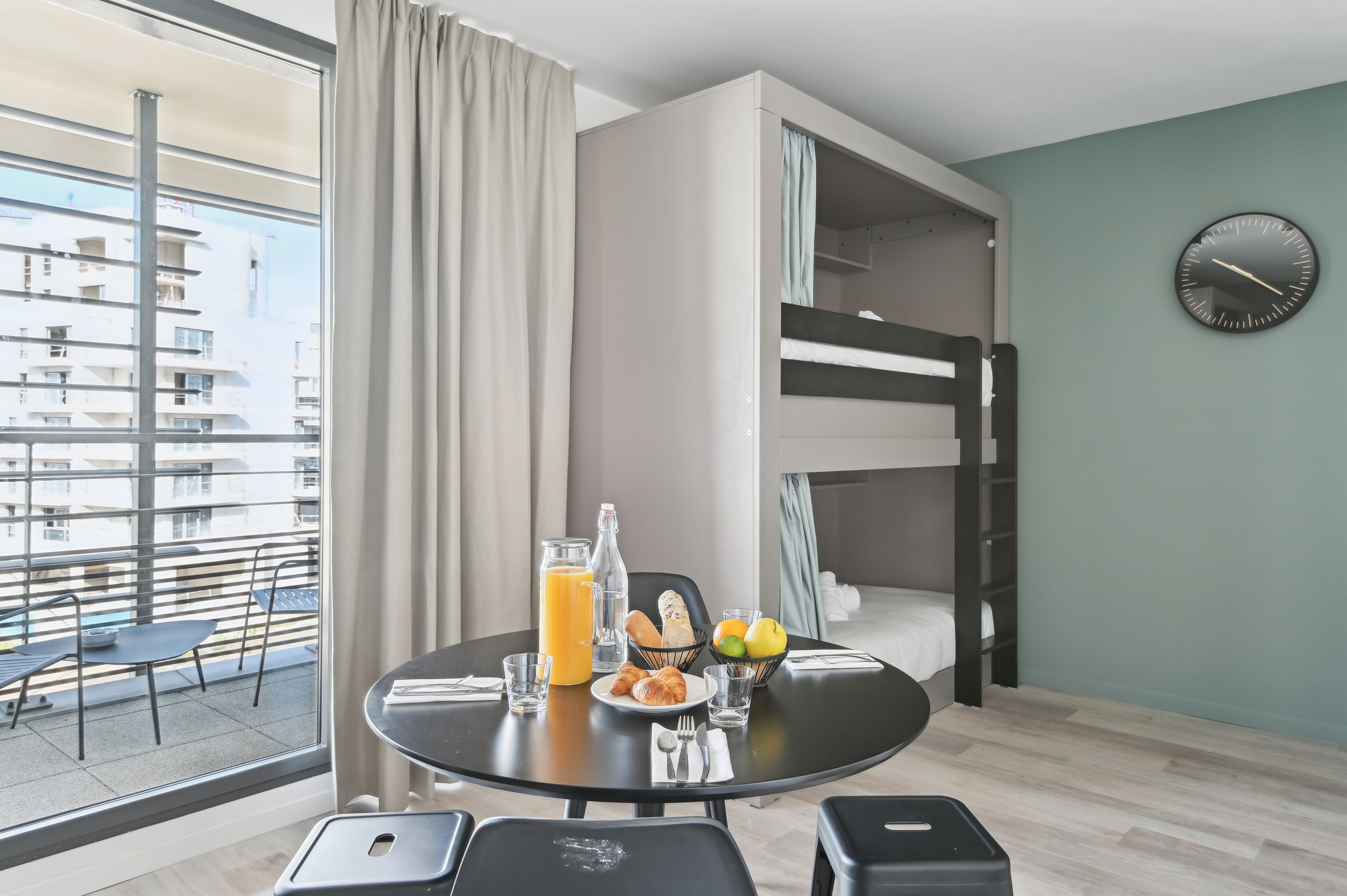 Une chambre d’hôtel moderne avec balcon, un lit superposé, un coin pour prendre le petit-déjeuner et une vue sur le paysage urbain.