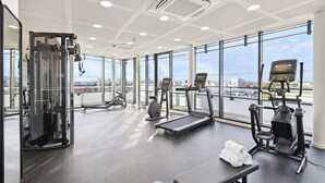A modern gym with various exercise equipment, large windows, and a city view. - All Suites Appart Hôtel | Paris Ouest - Colombes (Colombes)