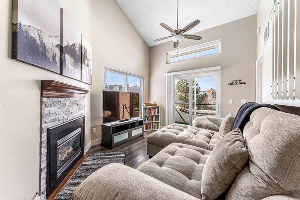 A living room with a fireplace, a large flat-screen TV, a ceiling fan, and a view of a cityscape through large windows.