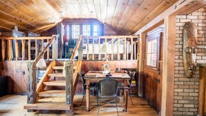 Interior rústico de madera con una escalera que conduce a un dormitorio en lo alto, un comedor con mesa y sillas, y una pared de ladrillo con una máscara de madera tallada.