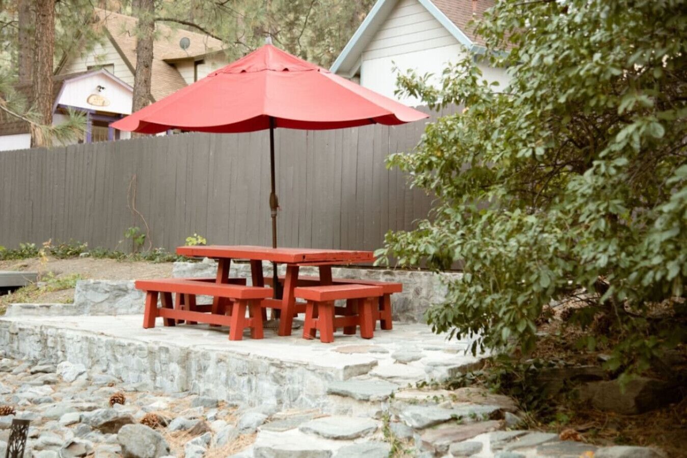 A red picnic table with an umbrella, set on stone steps in a backyard with a wooden fence and a house in the background.