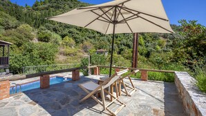 An outdoor pool area with lounge chairs and a large umbrella, surrounded by stone flooring and greenery.