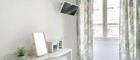 Une pièce lumineuse avec un bureau blanc, une petite plante, un tableau encadré et un téléviseur fixé au mur.