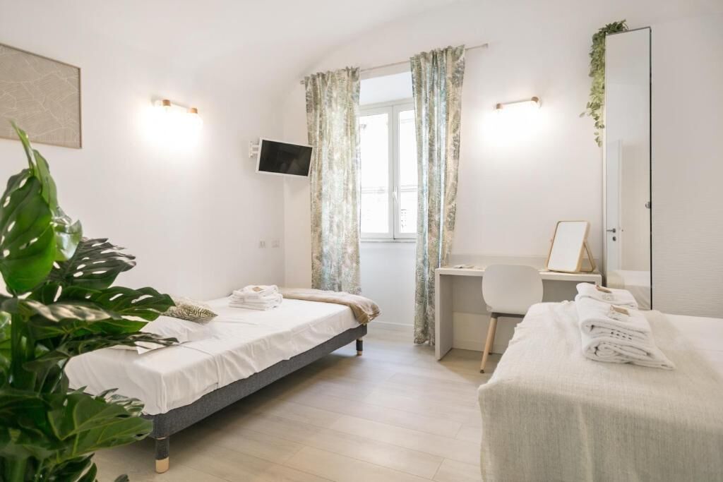 A hotel room with a bed, a desk, and a chair. There is a TV mounted on the wall, a window with curtains, and a plant.