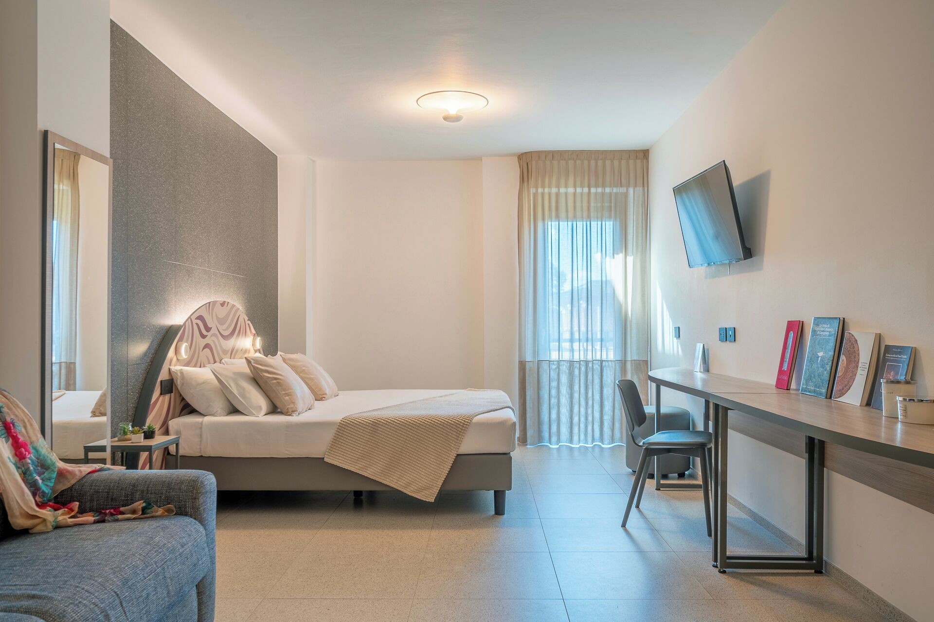A modern hotel room with a bed, bedside table, chair, desk, and a TV mounted on the wall.
